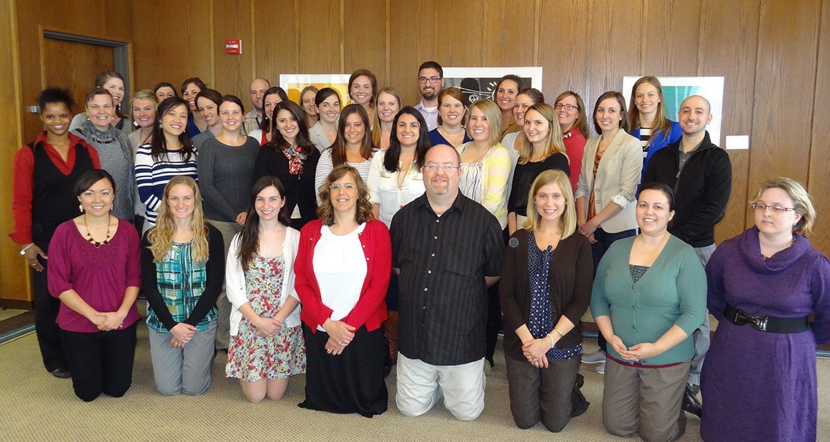 Cohort photo of 2012-2013 LEND trainees kneeling and standing in a room with wooden boarded walls.