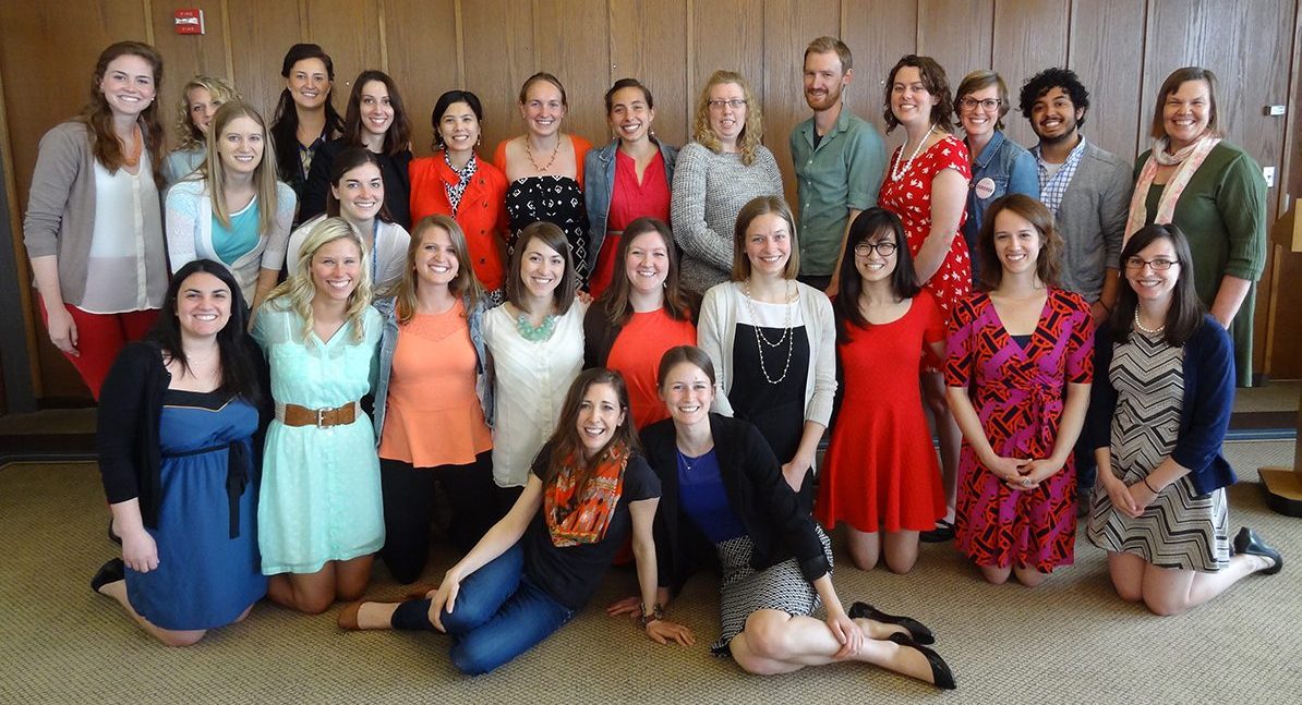 Cohort photo of 2013-2014 LEND trainees kneeling, sitting, and standing in a room with wooden boarded walls.
