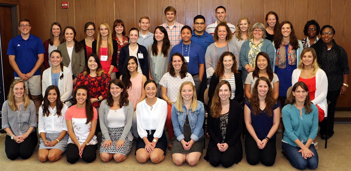 Cohort photo of 2017-2018 LEND trainees kneeling, sitting, and standing in a room with wooden boarded walls.