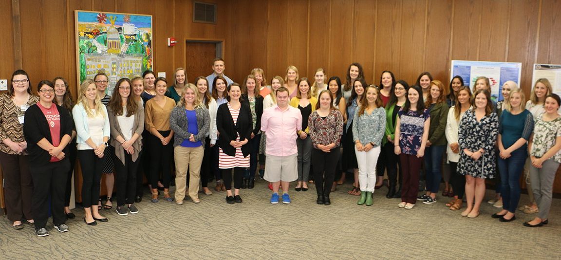 Cohort photo of 2018-2019 LEND trainees standing in a room with wooden boarded walls.