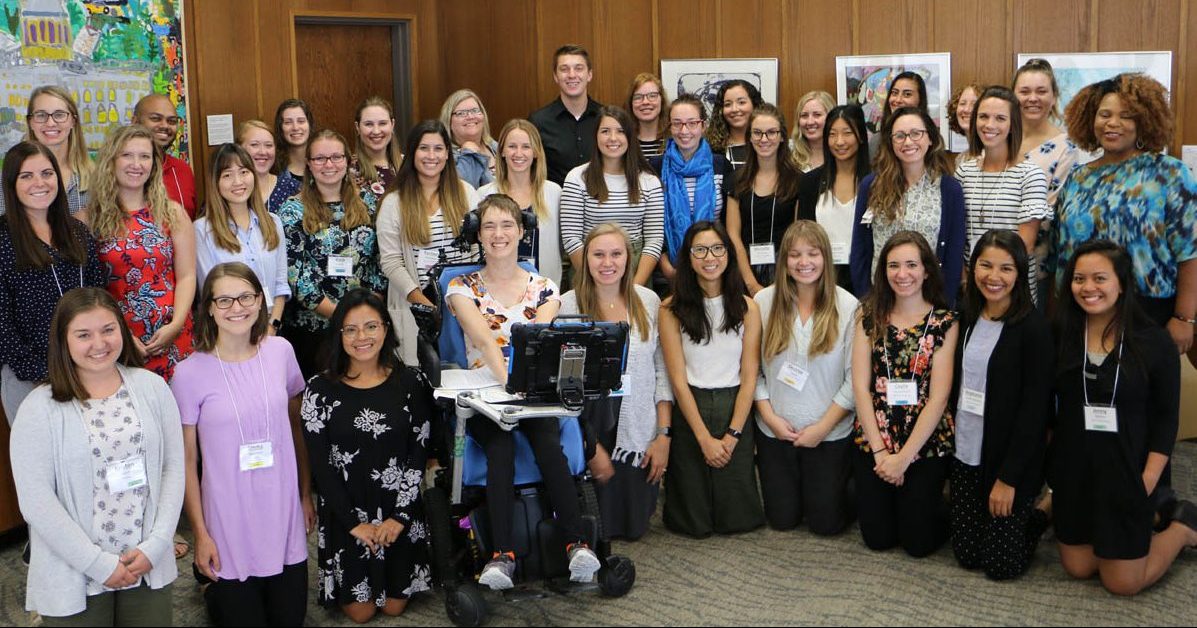 Cohort photo of 2019-2020 LEND trainees kneeling, sitting, and standing in a room with wooden boarded walls.