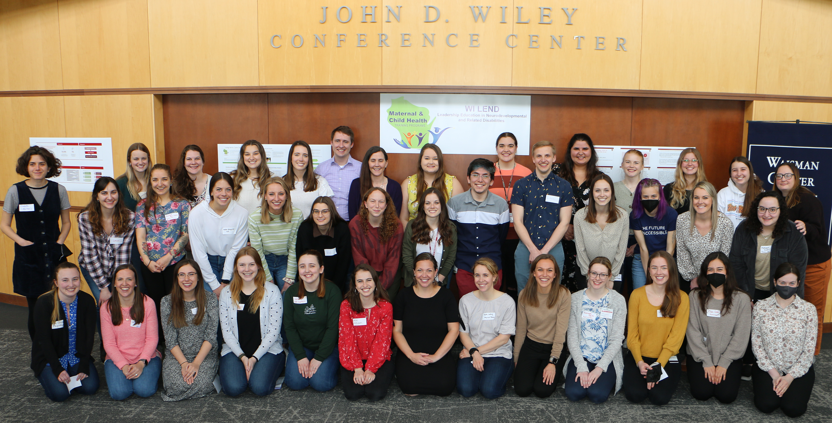 Cohort photo of 2021-2022 LEND trainees kneeling and standing in front of wooden paneled walls. Behind the trainees is a white banner with the WI LEND logo (green silhouette of Wisconsin with the words "Maternal and Child Health" on it along with icons of people. The banner also says "WI LEND" and "Wisconsin Leadership Education in Neurodevelopmental and Related Disabilities" in dark blue font.