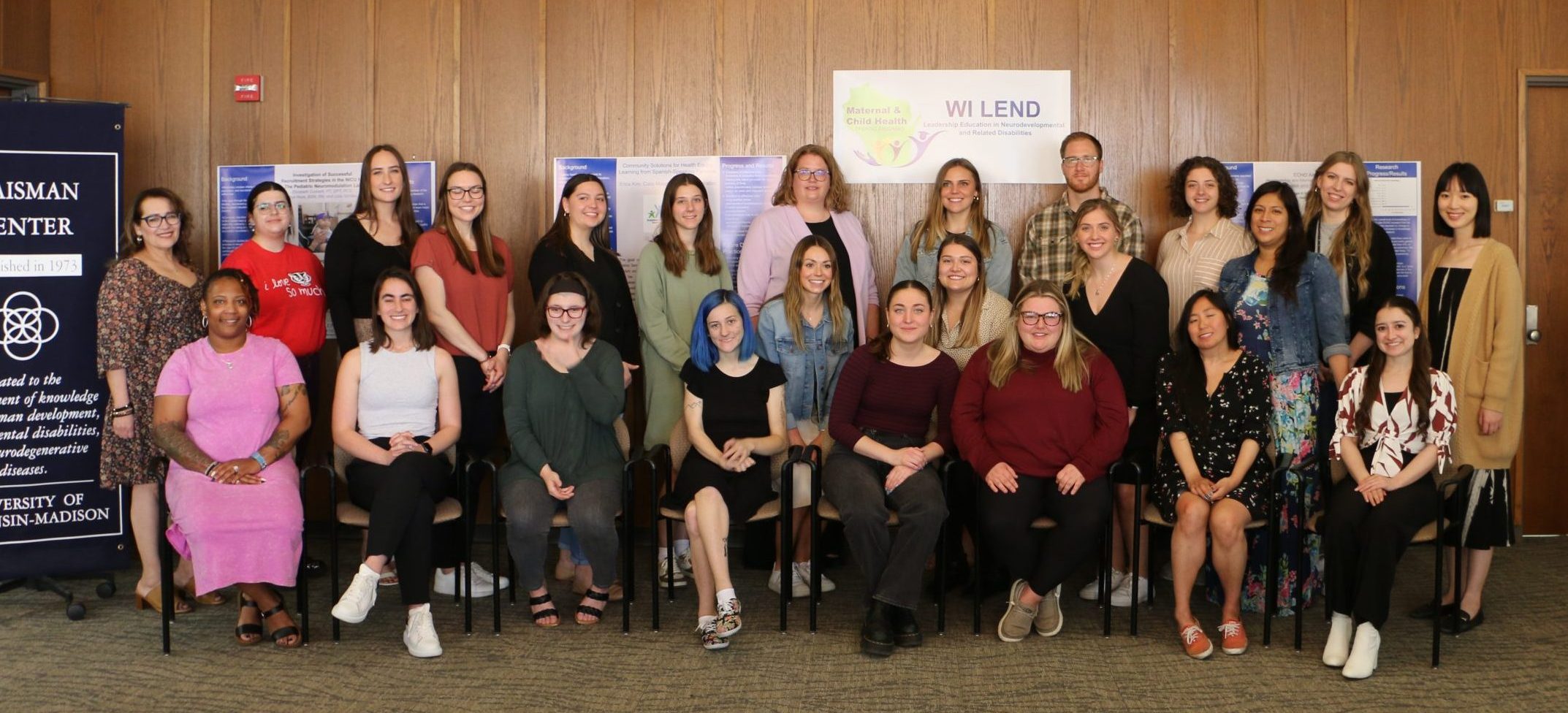 Cohort photo of 2022-2023 LEND trainees sitting and standing in a room with wooden boarded walls. Behind the trainees are blue and white posters, along with a white banner with the WI LEND logo (green silhouette of Wisconsin with the words "Maternal and Child Health" on it along with icons of people. The banner also says "WI LEND" and "Wisconsin Leadership Education in Neurodevelopmental and Related Disabilities" in dark blue font. To the left of the trainees is part of a navy blue Waisman Center banner.