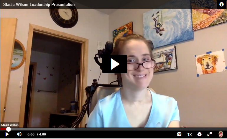 Video thumbnail depicting a young woman with CP in a power wheelchair with short red hair and purple glasses. She is wearing a light blue t shirt and is in a room with artwork on the walls. 