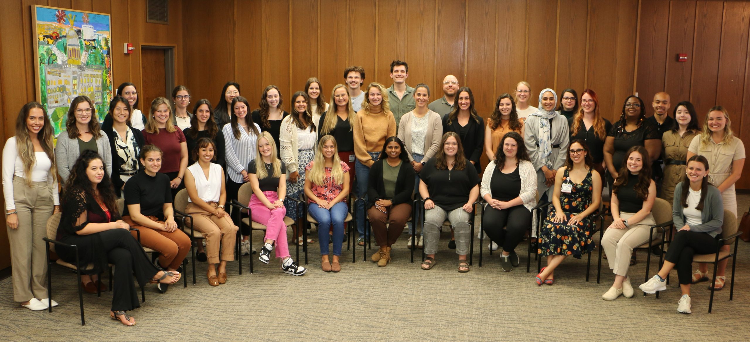 Cohort photo of 2023-2024 WI LEND trainees sitting and standing in a room with wooden boarded walls.
