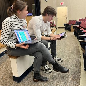 Young white woman holding a laptop with AAC device wearing a black and white striped top, grey jeans, and black boots. Sitting next to a young white man looking at tablet with AAC on it, wearing a white top, grey jeans, and sneakers. Sitting in the Waisman auditorium.