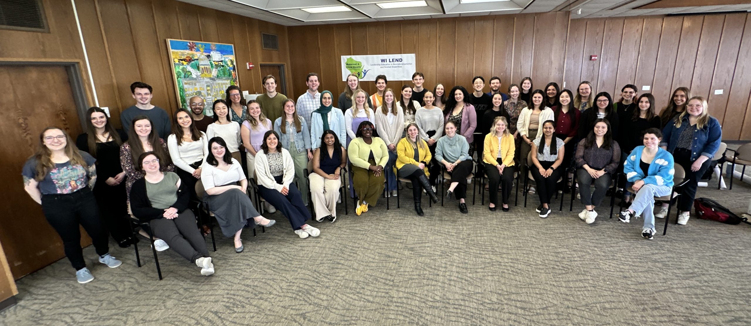 Cohort photo of 2024-2025 LEND trainees sitting and standing in a room with wooden boarded walls. Behind the trainees is a white banner with the WI LEND logo (green silhouette of Wisconsin with the words "Maternal and Child Health" on it along with icons of people. The banner also says "WI LEND" and "Wisconsin Leadership Education in Neurodevelopmental and Related Disabilities" in dark blue font.
