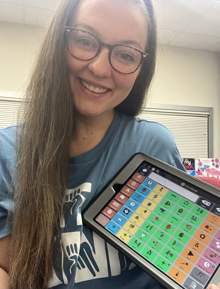 Photo of young woman with long brown hair and glasses holding an AAC device with colored columns.