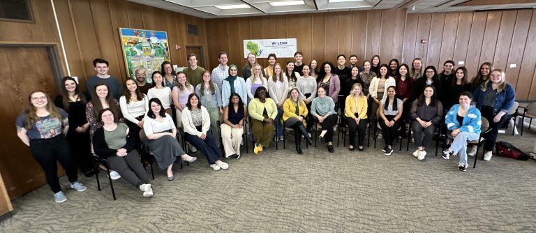 Cohort photo of 2024-2025 LEND trainees sitting and standing in a room with wooden boarded walls. Behind the trainees is a white banner with the WI LEND logo (green silhouette of Wisconsin with the words "Maternal and Child Health" on it along with icons of people. The banner also says "WI LEND" and "Wisconsin Leadership Education in Neurodevelopmental and Related Disabilities" in dark blue font.