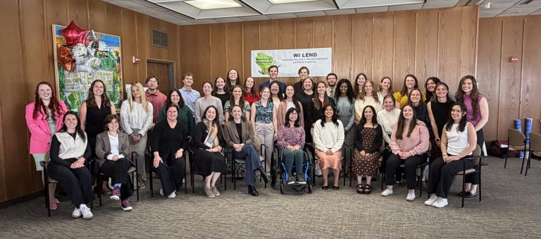 Cohort photo of 2025-2026 LEND trainees sitting and standing in a room with wooden boarded walls. Behind the trainees is a white banner with the WI LEND logo (green silhouette of Wisconsin with the words "Maternal and Child Health" on it along with icons of people. The banner also says "WI LEND" and "Wisconsin Leadership Education in Neurodevelopmental and Related Disabilities" in dark blue font.