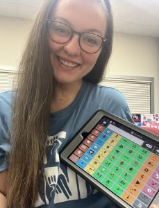 Photo of young woman with long brown hair and glasses holding an AAC device with colored columns.
